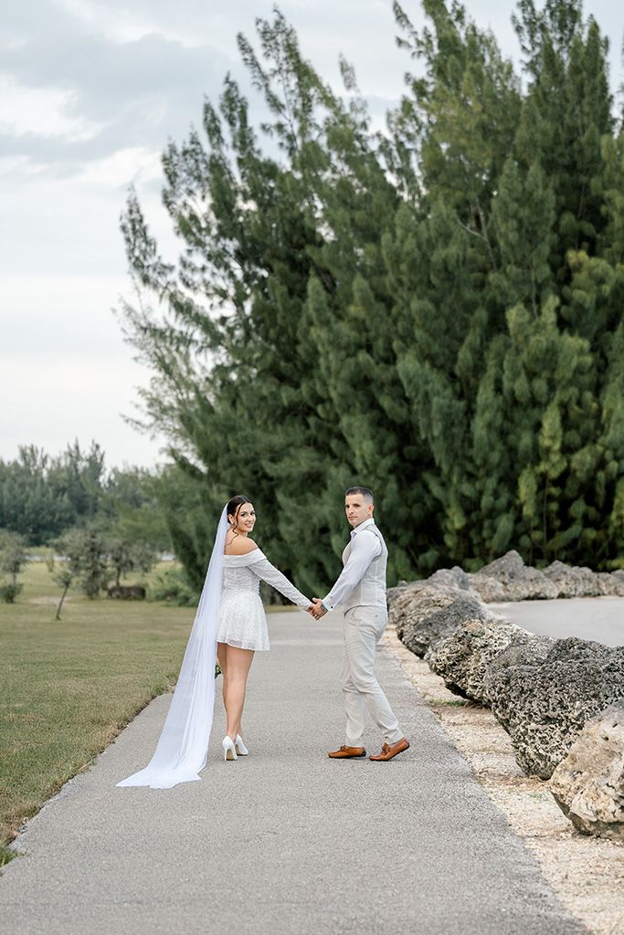 Bride and groom hold hands on a path surrounded by tall green trees, bride wearing mini dress with long cathedral veil.