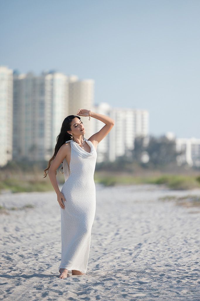 Woman in white maxi dress posing on sandy beach with coastal buildings in background on a clear day.