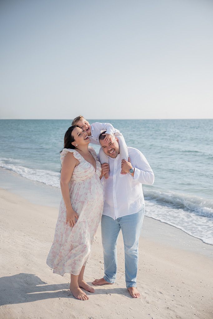 Happy family laughing together on a sunny beach, woman in floral dress and man in white shirt carrying child.