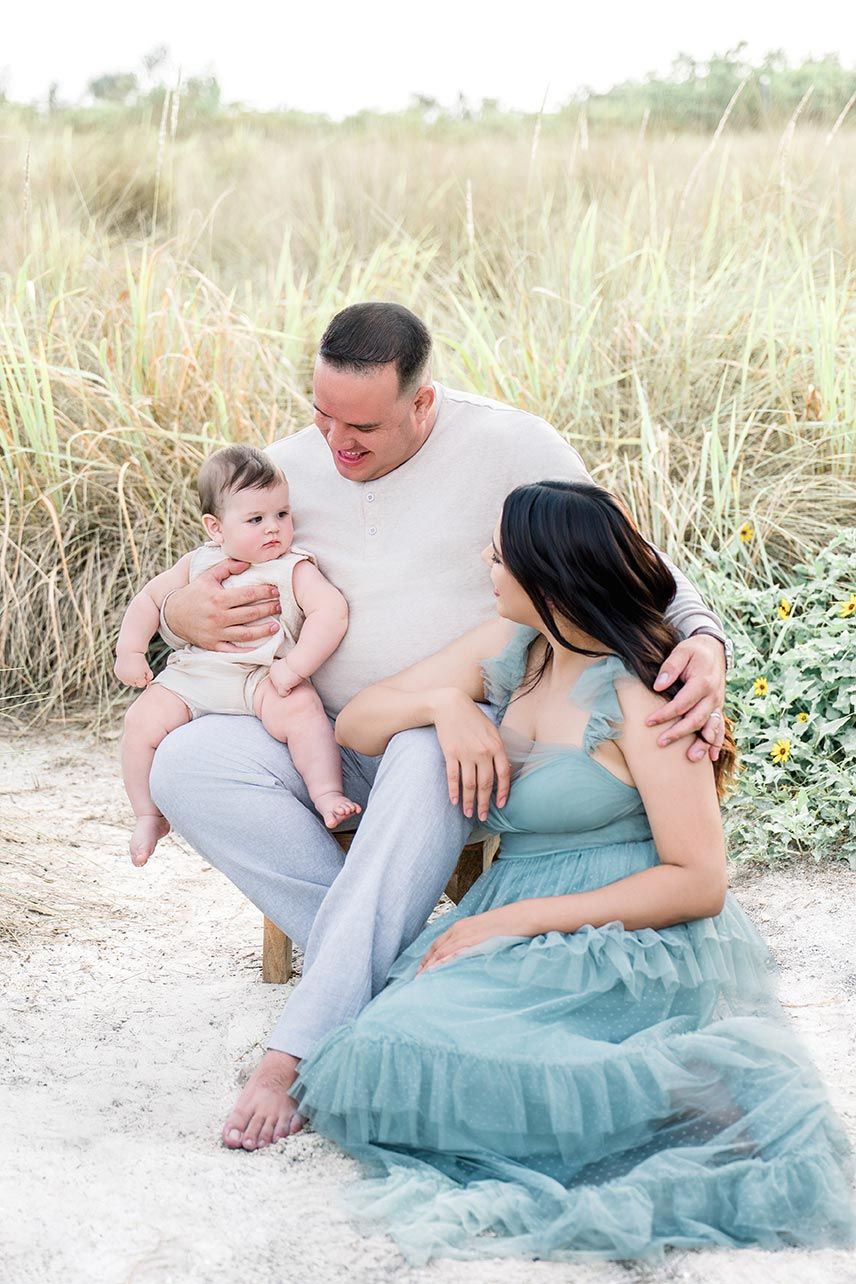 A family of three poses on sandy beach dunes, father holding baby while mother in teal tulle dress sits beside them.