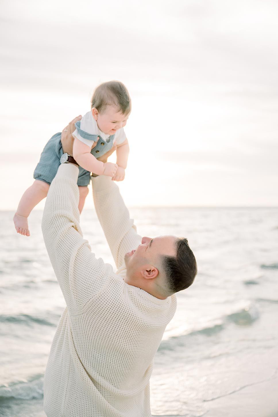 A father lifts his smiling baby high in the air on a serene beach at sunset, both dressed in light blue and white.