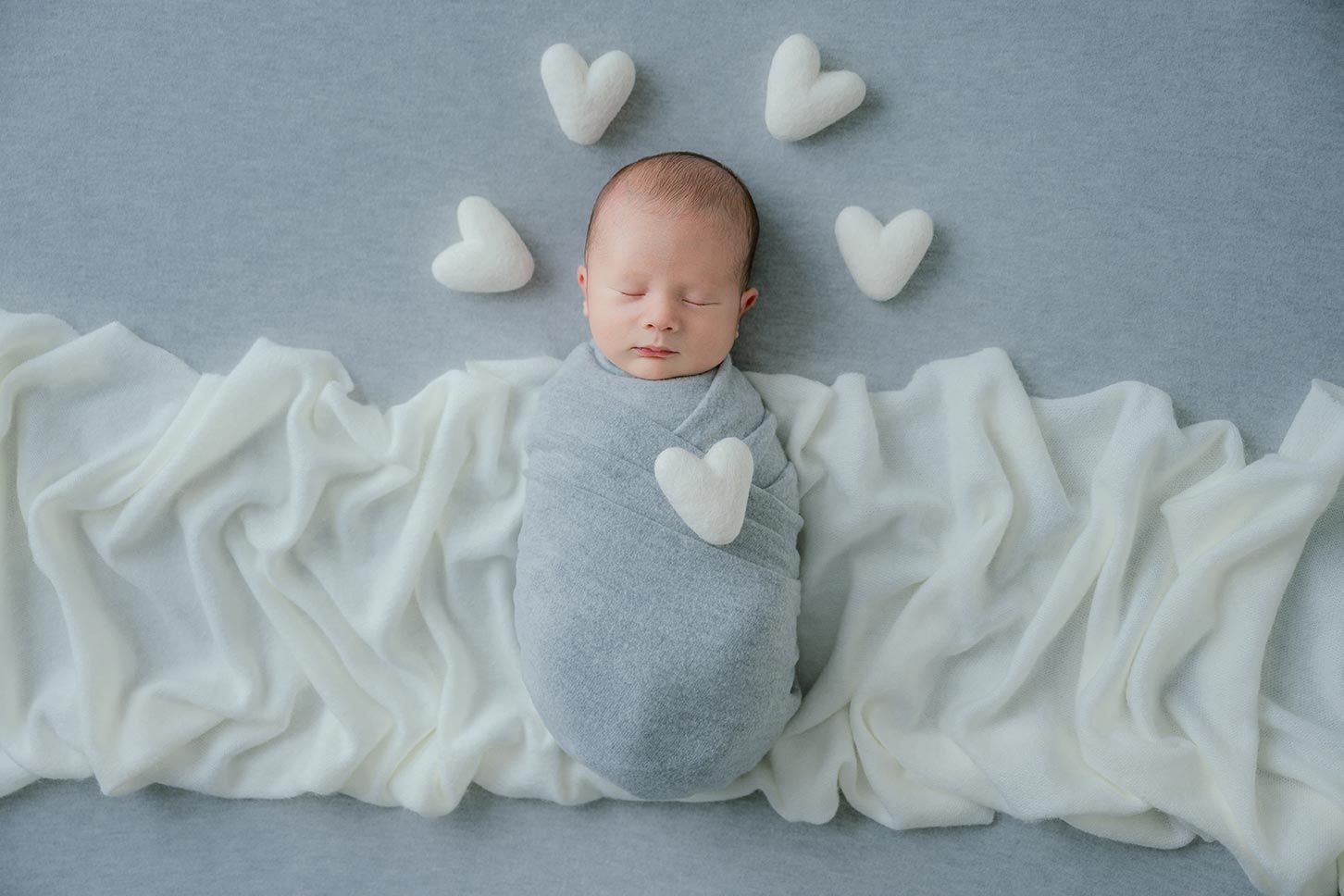 Newborn baby swaddled in gray wrap surrounded by white heart props on a soft blue backdrop.