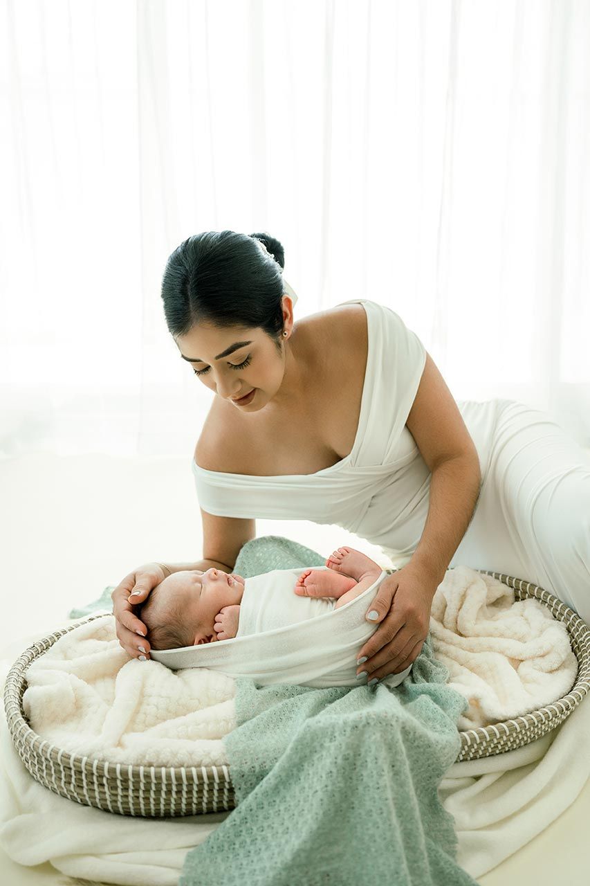 Mother in white dress lovingly gazing at her swaddled newborn baby lying in a wicker basket.
