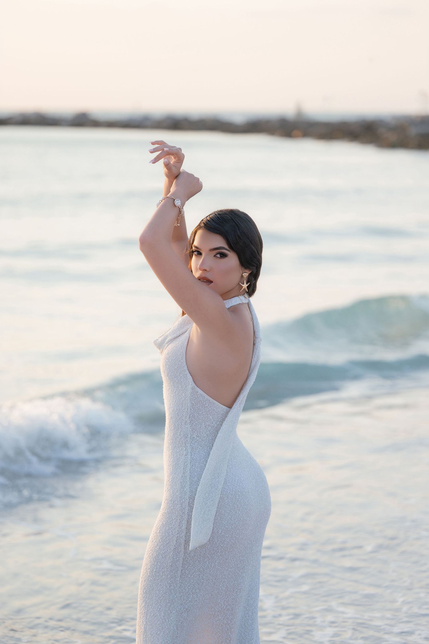 Woman in white backless gown poses gracefully on beach at sunset with waves behind her.
