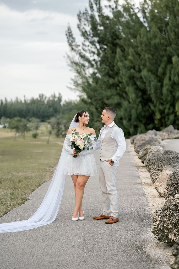Bride in short white dress and veil holds bouquet while gazing at groom in beige suit on scenic outdoor path.
