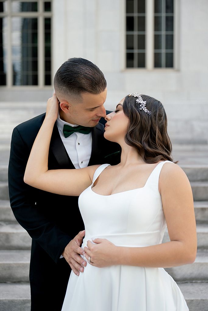 Bride and groom share a romantic almost-kiss on stone steps, she in white gown, he in black tuxedo with green bow tie.