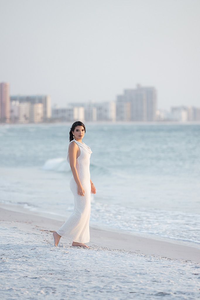 Woman in flowing white gown walking barefoot along ocean shoreline with city skyline.