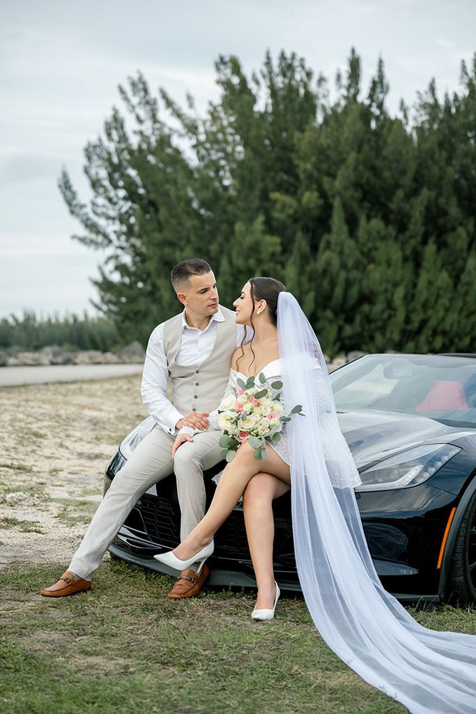 Bride and groom pose together on a black sports car hood outdoors, with tall green trees in the background.