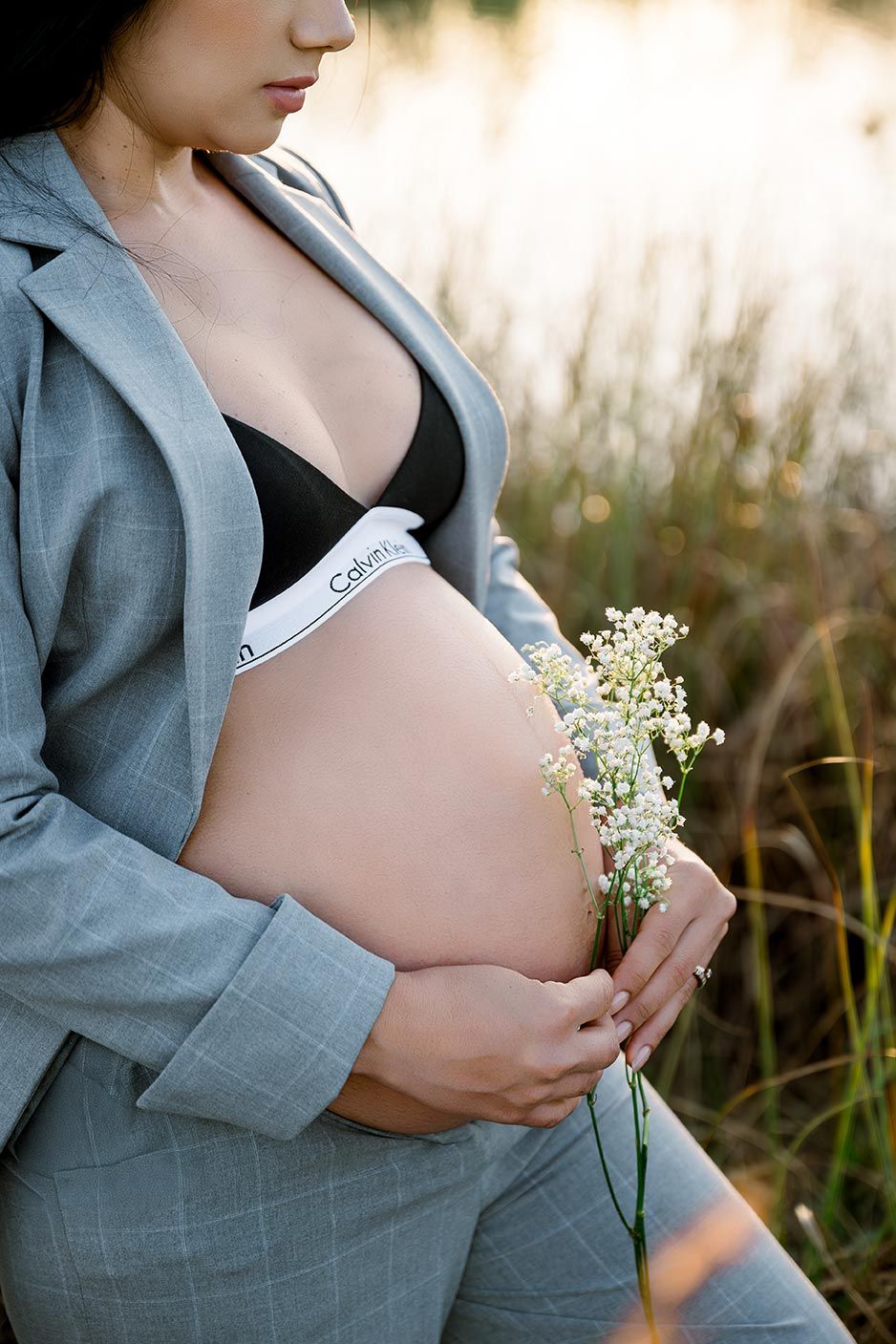 A pregnant woman in a gray blazer holds delicate white flowers against her belly in a golden-lit outdoor setting.