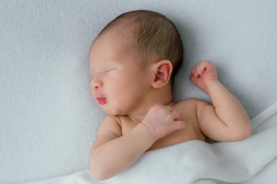 A sleeping newborn baby rests peacefully against soft white fabric with arm outstretched.