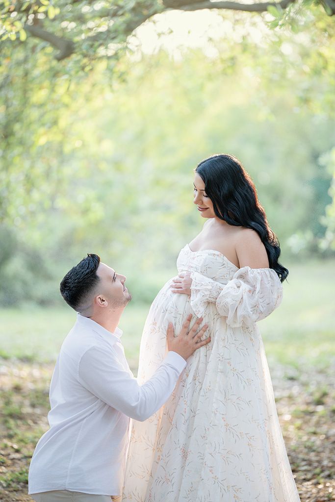 Man kneels before pregnant woman in floral lace gown, both smiling in a sunlit park setting.