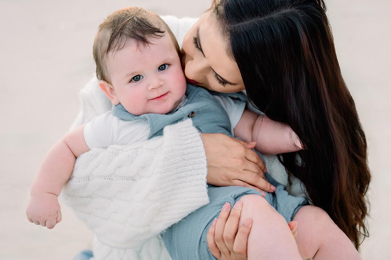 A dark-haired mother tenderly kisses her smiling baby dressed in a blue outfit on a sunny beach.