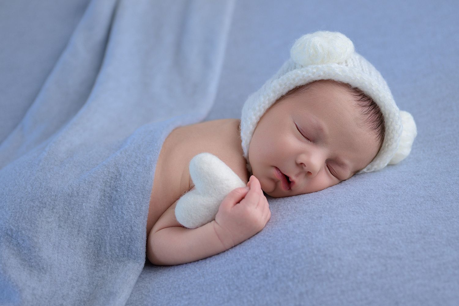 Newborn baby sleeping peacefully on blue blanket, wearing white knit bear hat, holding small white heart toy.