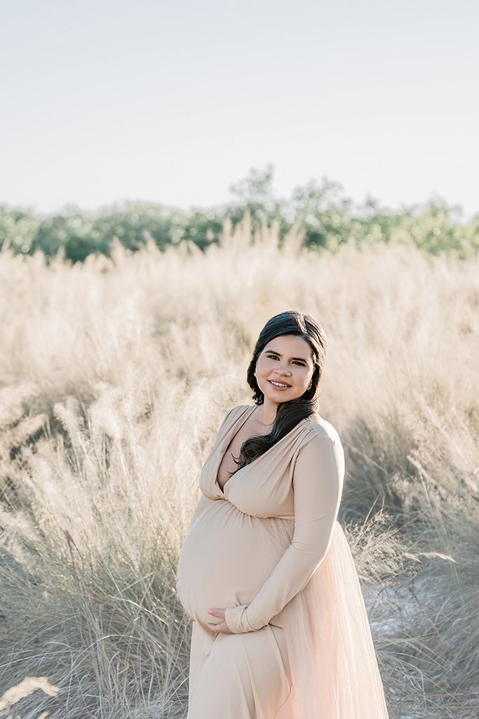 Smiling pregnant woman in a blush maxi dress cradling her baby bump in a golden field of tall grass.