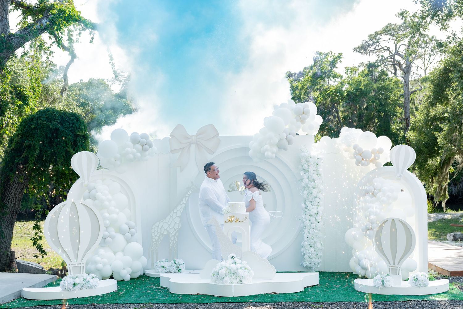 Couple in white attire poses at elegant outdoor wedding backdrop with white balloons, floral decor, and blue smoke.