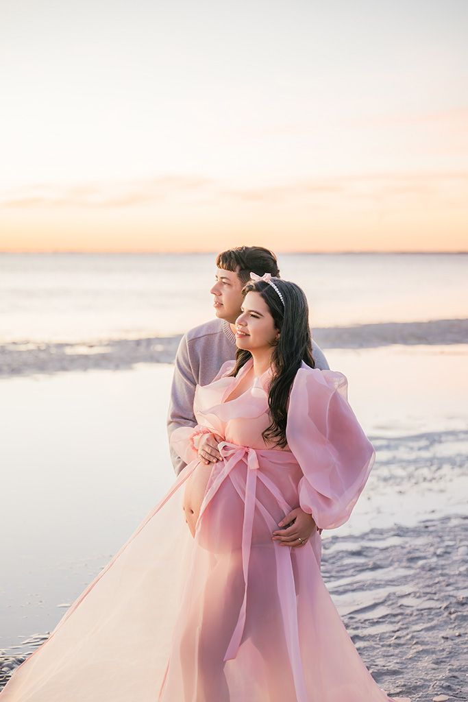 Couple embracing at sunset on the beach, woman in flowing pink gown, soft pastel sky reflecting on calm water.