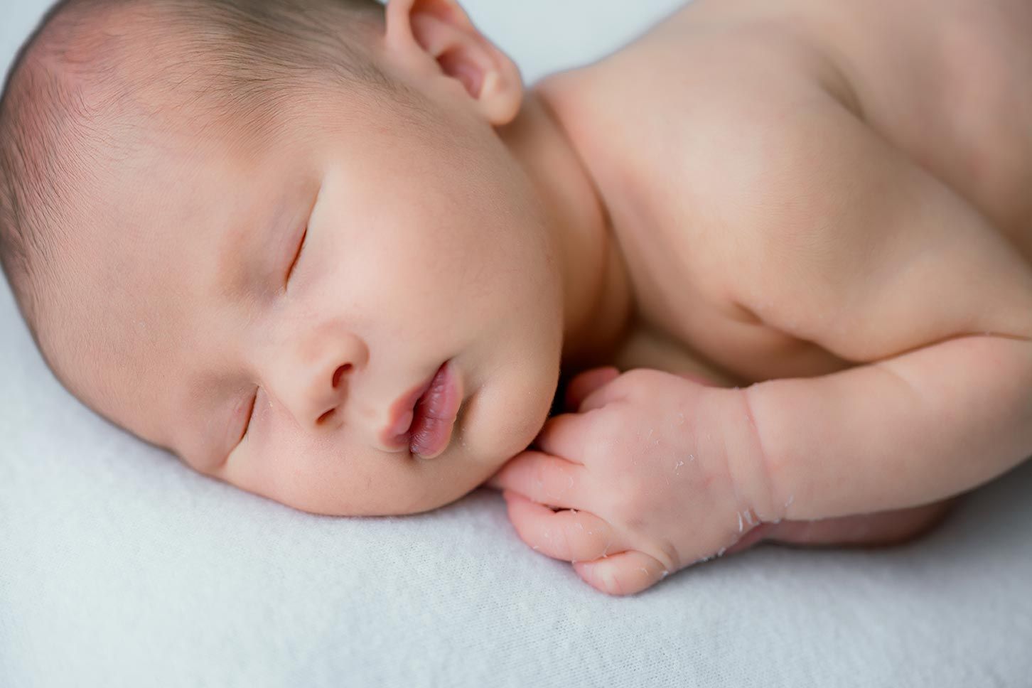 Peaceful newborn baby sleeping on white blanket with tiny hands tucked under chin in serene portrait.