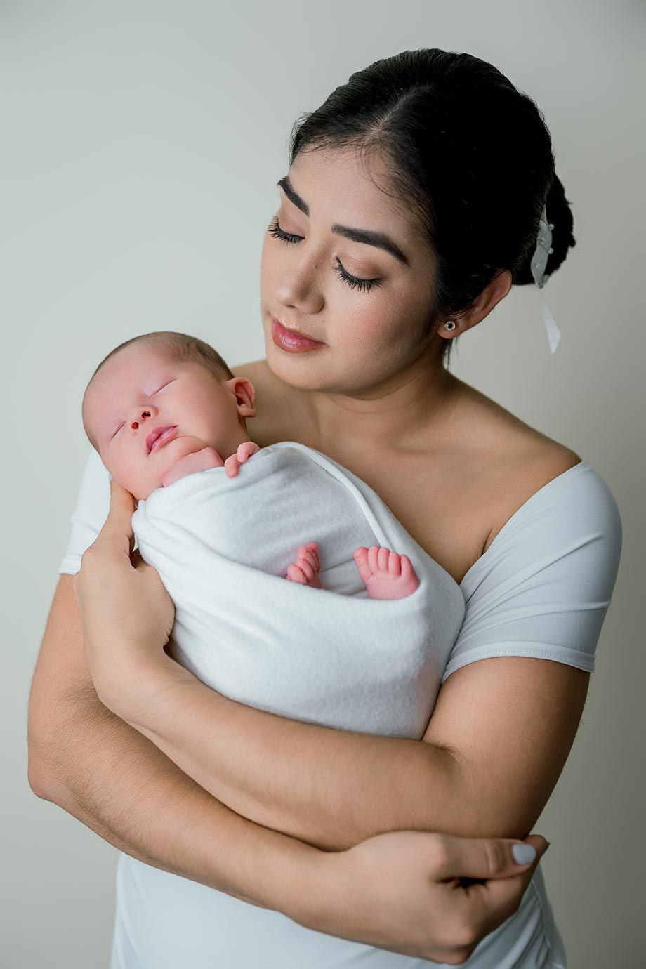 Young mother in white off-shoulder top tenderly holding her sleeping swaddled newborn baby.