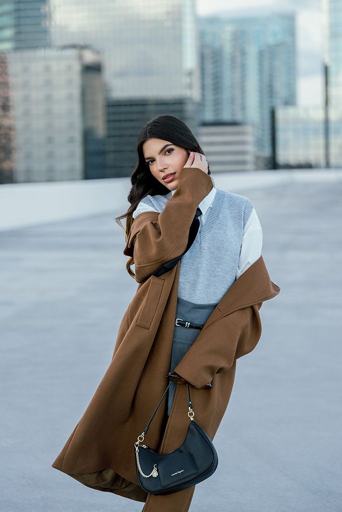 Woman in brown longline coat with grey vest and black crossbody bag poses on urban rooftop.