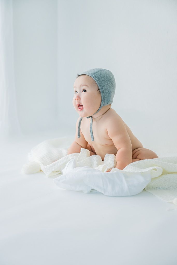 Cute baby wearing a gray bonnet hat sitting on white blankets in a bright white studio setting.