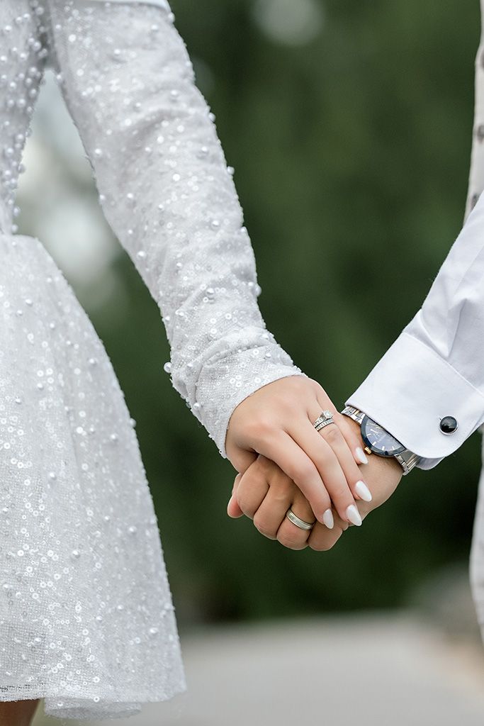 Close-up of newlyweds holding hands, bride wearing sparkly white dress and wedding ring, groom in white shirt with watch.