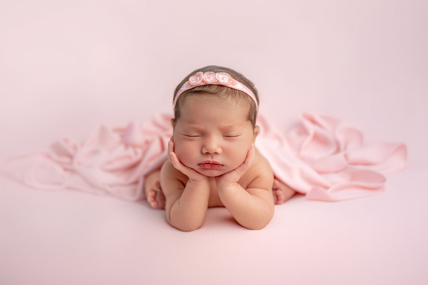 Sleeping newborn baby girl in pink wrap and floral headband posed with hands under chin on pink background.