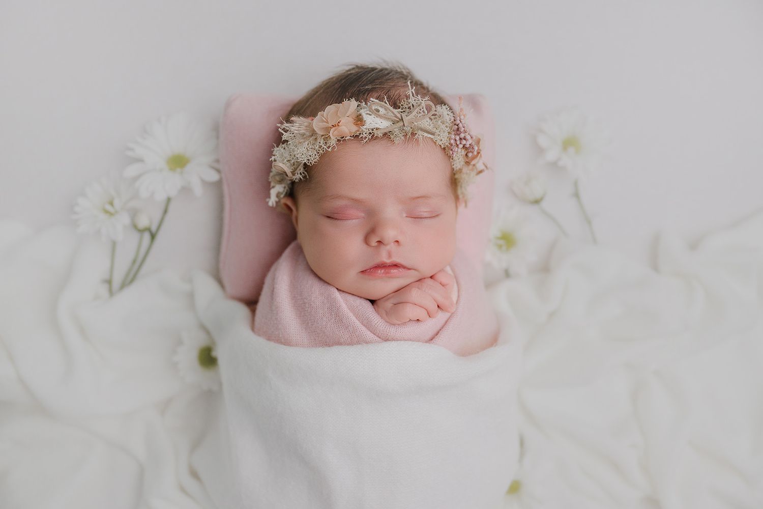Sleeping newborn baby girl wrapped in pink swaddle with floral headband, surrounded by white daisies on white background.