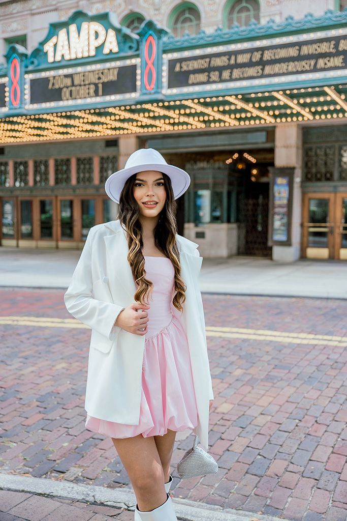 Stylish woman wearing pink dress, white blazer and hat standing before Tampa Theatre marquee sign.