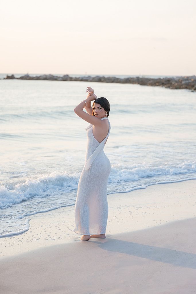 Woman in white dress standing on beach with ocean waves, arms raised, at sunset.