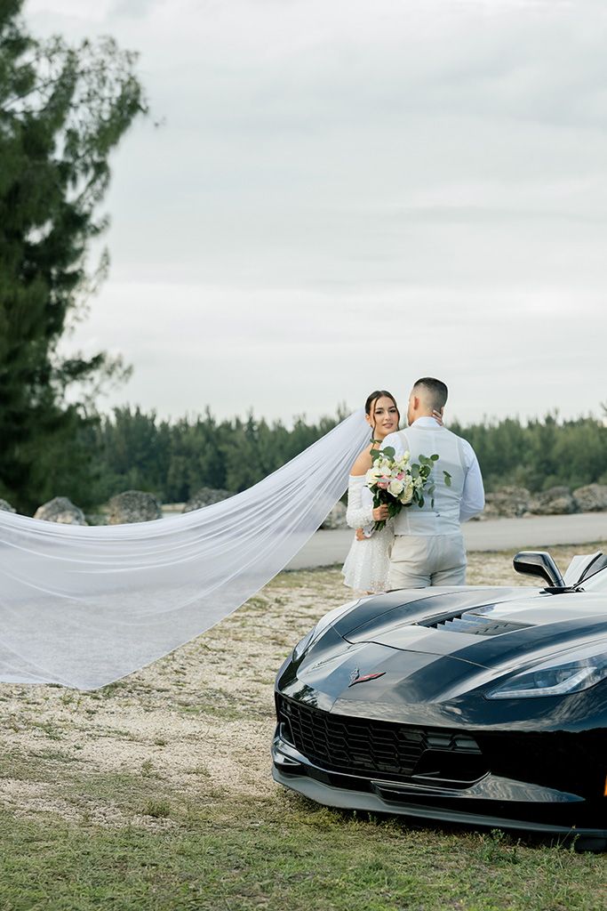 Bride and groom embrace near black Corvette on sandy shore, her long veil flowing in the breeze.
