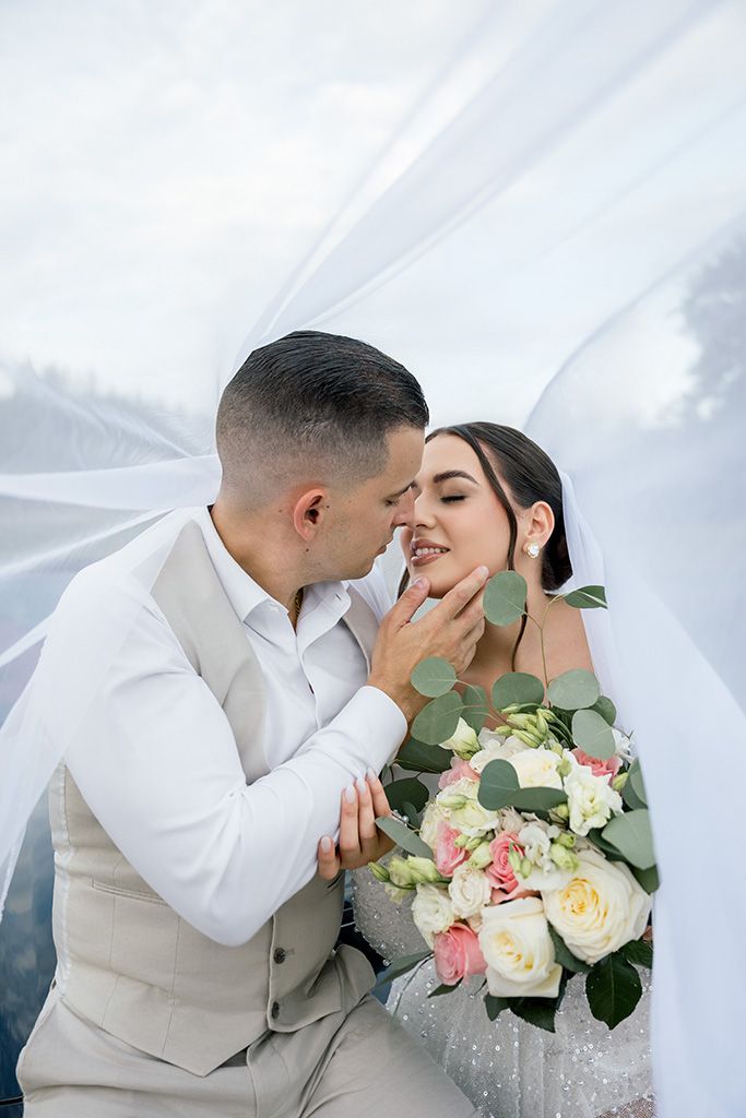 Bride and groom share a romantic moment under a white veil, bride holding a bouquet of pink and white roses.