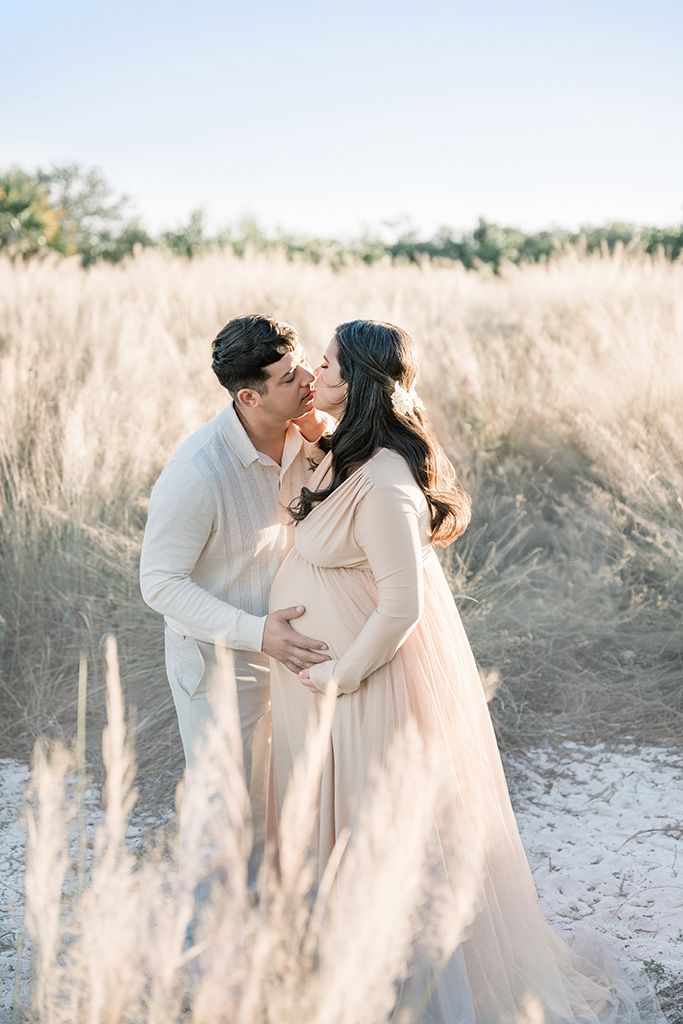 A couple shares a tender kiss outdoors, the pregnant woman in a flowing blush gown, surrounded by golden tall grasses.