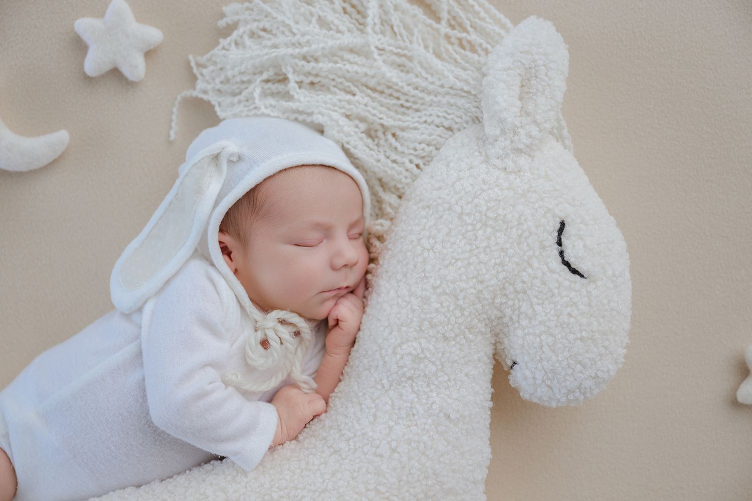 Close-up of a peaceful newborn baby in white hat sleeping against a soft fluffy unicorn stuffed animal.