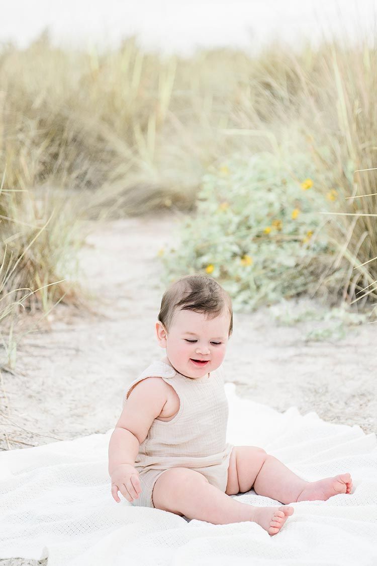 A baby wearing a white onesie sits and plays on a sandy beach with dunes and wildflowers in the background.