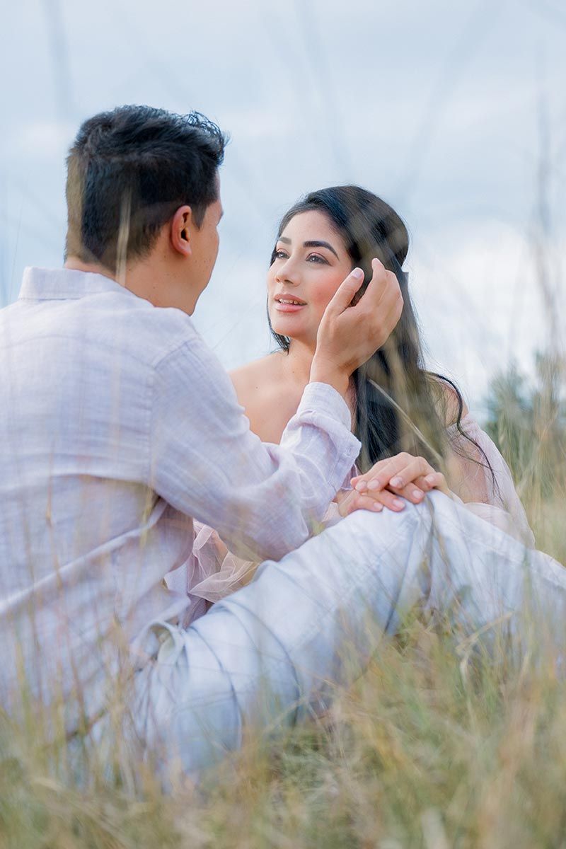 A romantic couple sits in tall grass wearing white outfits, sharing an intimate moment during a pre-wedding photoshoot.