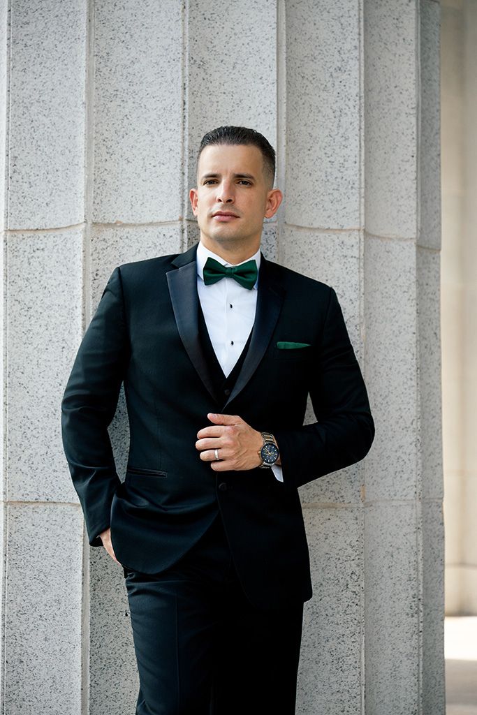 Man in black tuxedo with green bow tie and pocket square, posing confidently against stone columns.