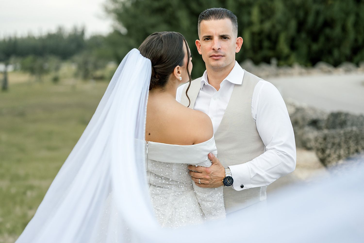 Groom in gray vest embraces bride in lace off-shoulder wedding dress with flowing veil outdoors.