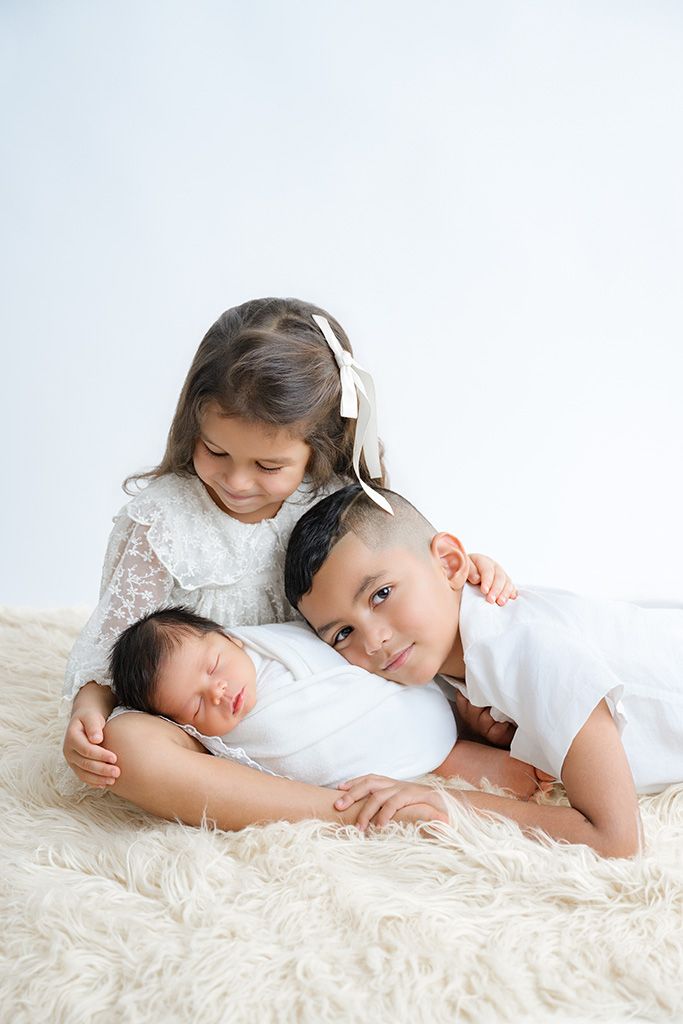 Two siblings tenderly pose with sleeping newborn baby on fluffy cream rug in white studio setting.