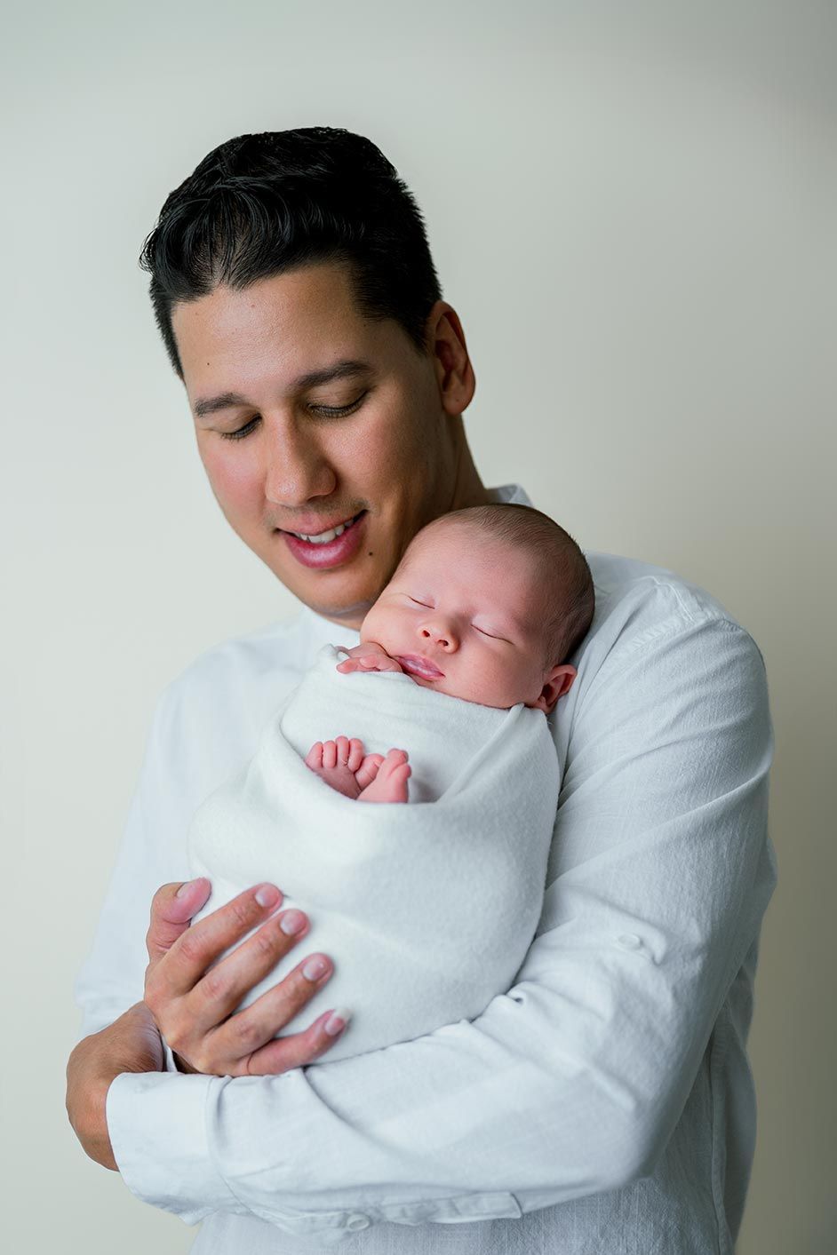 Young father in white shirt smiling while cradling sleeping swaddled newborn baby against his chest.