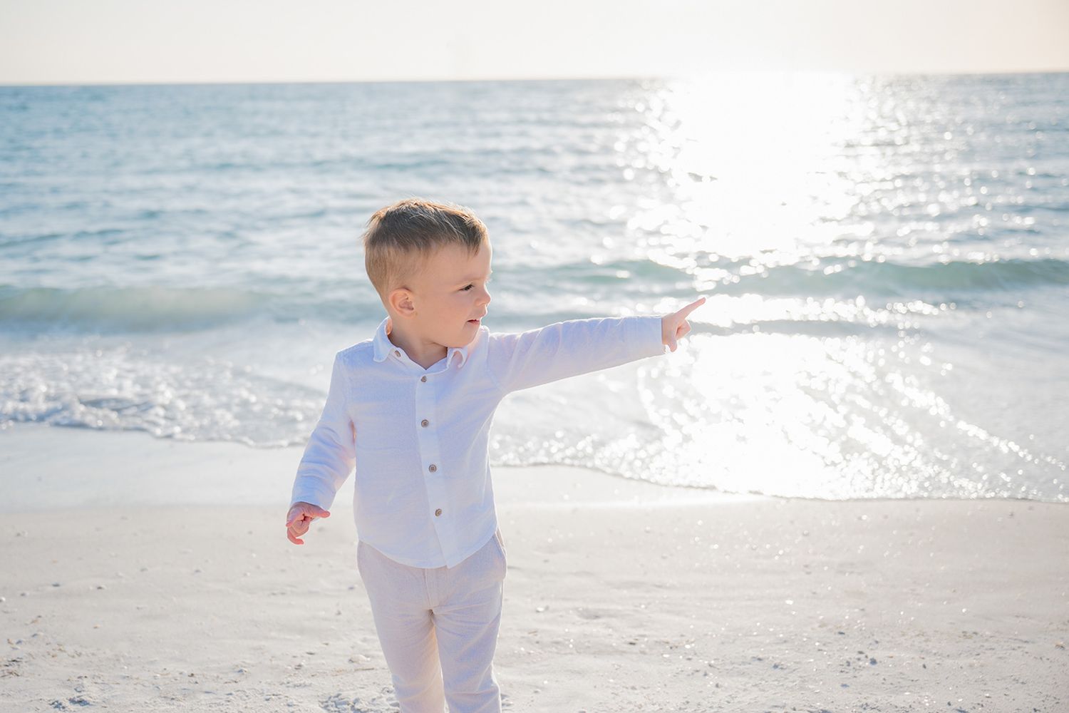 Young boy in white outfit pointing at the ocean on a sunny beach day.