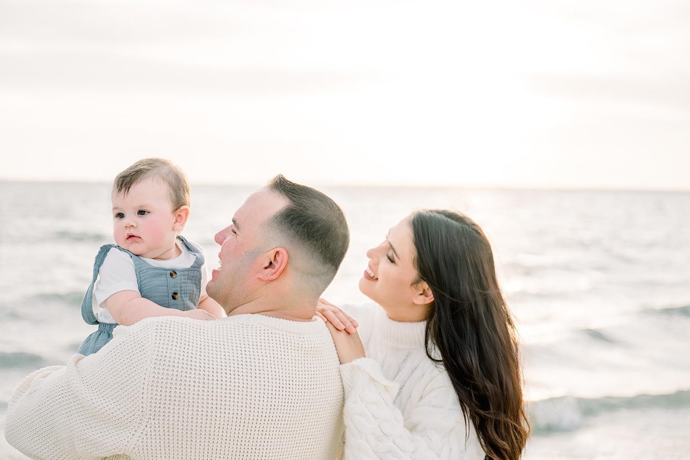 Family with baby boy at beach during golden hour, parents smiling lovingly at child by ocean.