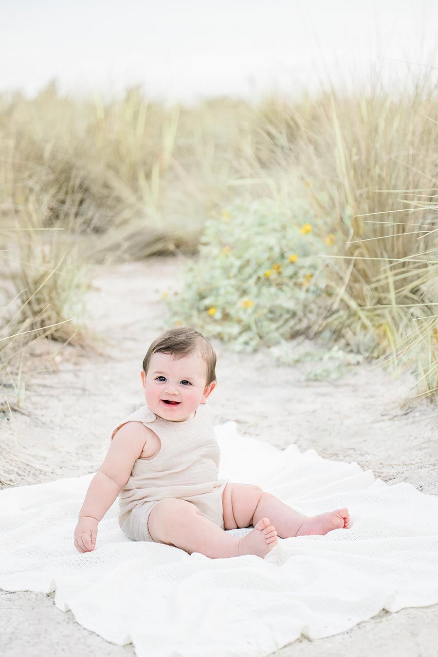 A happy baby sits on a white blanket among sandy beach dunes, wearing a beige romper, smiling at the camera.