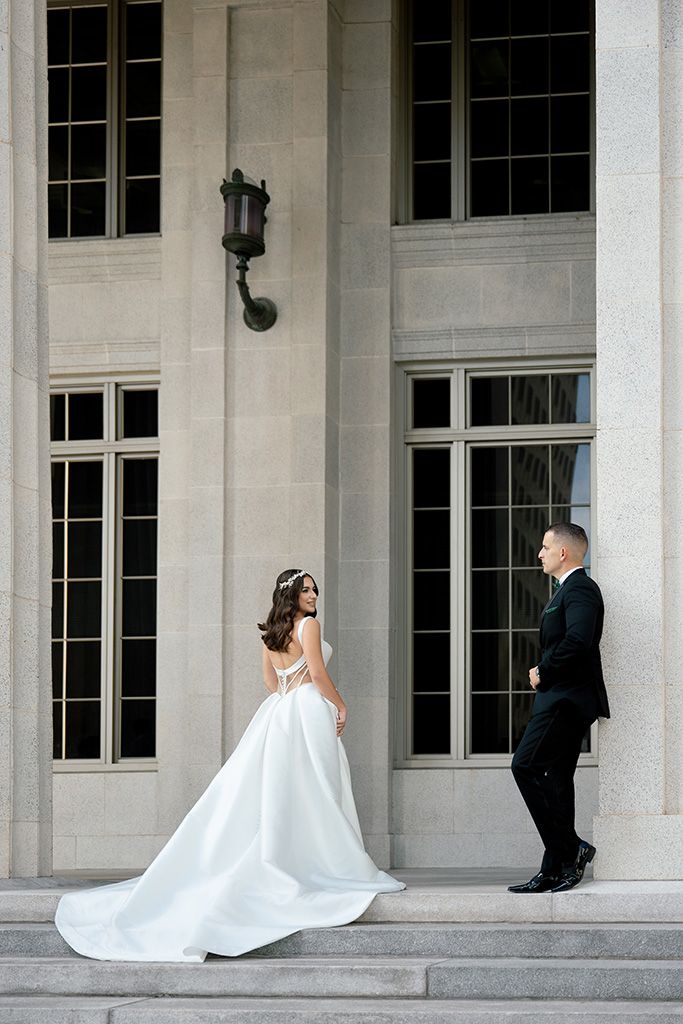 Bride in elegant white gown with train and groom in black tuxedo pose on grand stone steps of classic building.