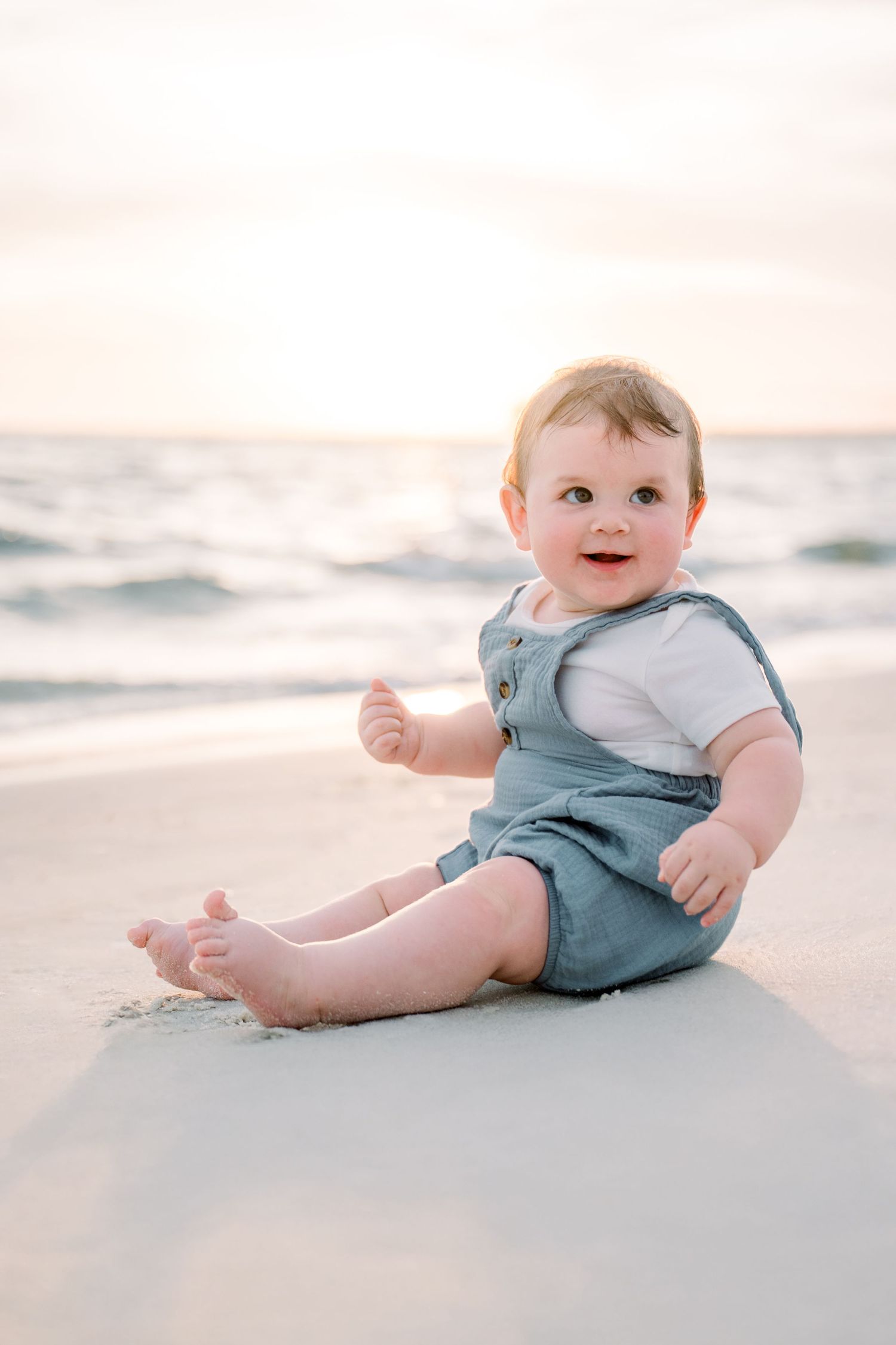 A baby in light blue overalls sits and plays on a pristine white sandy beach at sunset.
