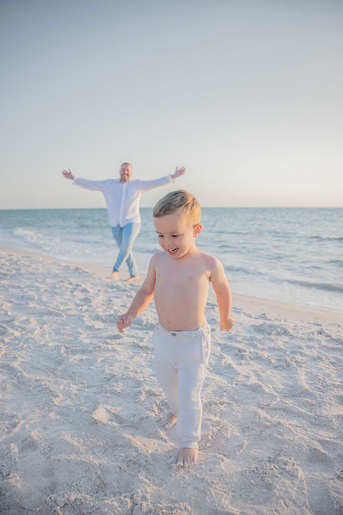 Happy shirtless toddler running on white sand beach with father posing arms wide near ocean shore.
