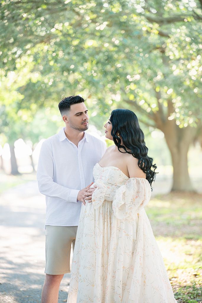A couple shares a tender moment on a tree-lined path, woman in lace maternity gown, man in white shirt and khaki shorts.