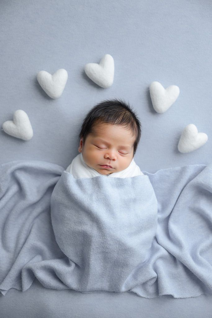 Sleeping newborn baby swaddled in a light blue blanket surrounded by white felt hearts on a soft gray background.