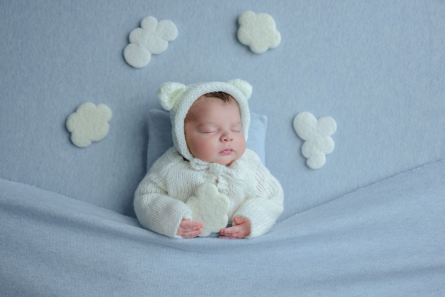 Newborn baby sleeping peacefully in a white bear outfit on a soft blue blanket with cloud decorations.