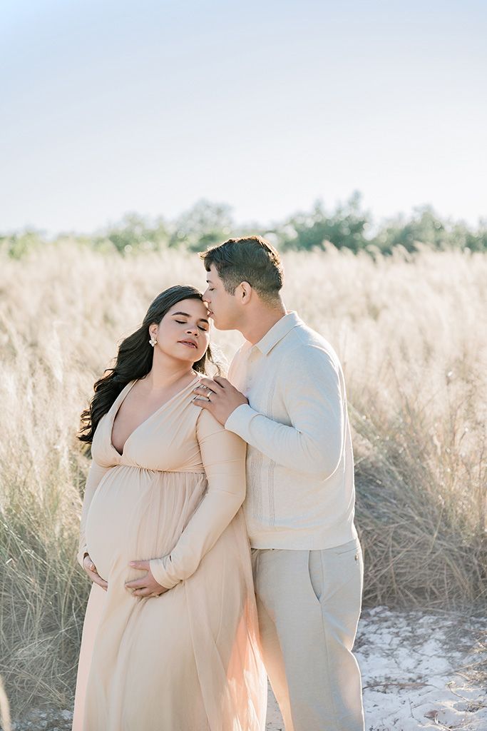 A couple shares a tender moment in a golden field, the man kissing his pregnant partner's temple as she cradles her belly.