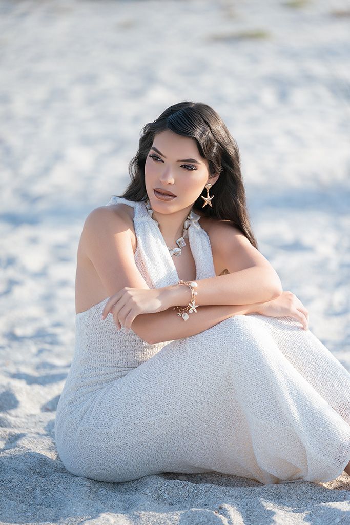 Young woman in white sequin dress with starfish jewelry sitting gracefully on sandy beach.